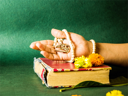 open hand showing Aum locket with beads, red hindu book in the background with flowers