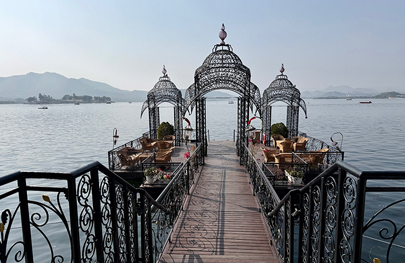 This scene depicts a pedestrian walkway and decorative iron arches leading to a pier or jetty on Lake Pichola in Udaipur, India, often used for accessing luxury hotels like the Taj Lake Palace. 