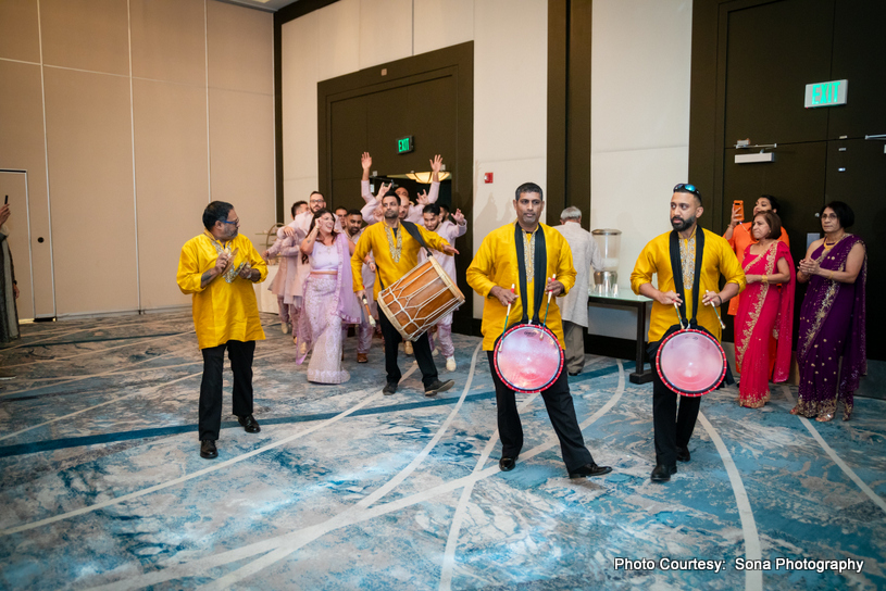 Traditional Indian wedding celebration with drummers and guests in festive attire enjoying music and dance indoors.