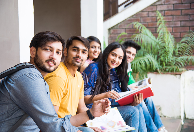 Young Asian Indian college students reading books, studying on laptop, preparing for exam or working on group project while sitting on grass, staircase or steps of college campus